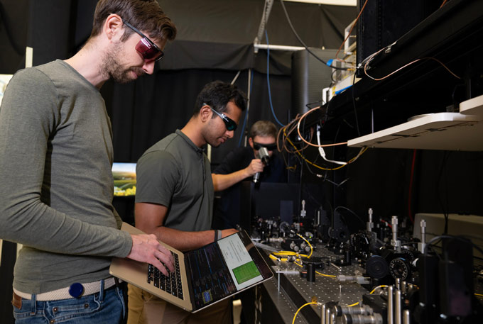 Três homens em um laboratório, um em primeiro plano segurando um laptop aberto, estão trabalhando em um projeto para gerar números aleatórios.